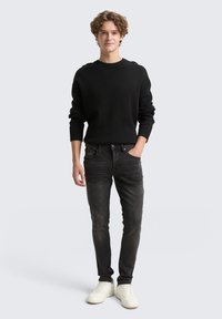 Čierny pletený sveter, priliehavý, s okrúhlym výstrihom; tmavošedé slim-fit džínsy; biele tenisky s textúrovanými stranami; model stojí s jednou rukou v vrecku.