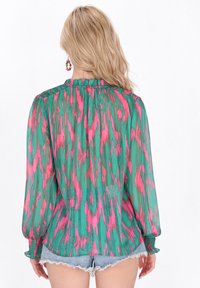 Blusa verde trasparente con motivo astratto rosa e teal. Presenta scollatura arricciata e maniche lunghe con polsini elasticizzati, abbinata a pantaloni shorts in denim.