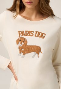 Top de manga larga beige con el texto "PARIS DOG" y un gráfico bordado de un perro marrón con un acabado texturizado y acentos plateados.