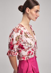Blumenbluse mit pinkem Hintergrund, versehen mit roten und goldenen Blumen, gepufften Ärmeln und einem tiefen V-Ausschnitt, kombiniert mit leuchtend pinken Hosen.