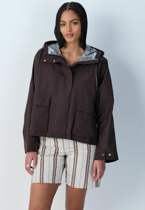 Frau trägt eine dunkle Kapuzenjacke mit großen Fronttaschen und gestreifte beiger Shorts, steht vor einem einfarbigen hellen Hintergrund.