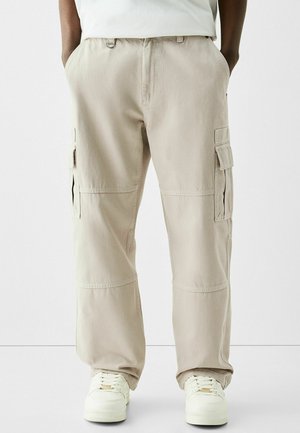 Person iført beige cargobukser med flere lommer og hvide sneakers, stående med hænderne i lommerne mod en ensfarvet baggrund.
