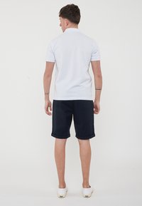 Polo shirt bianco a maniche corte, pantaloni corti blu navy con orli arrotolati e scarpe bianche senza lacci, viste da dietro. Design semplice e casual.