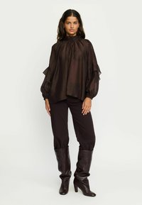 Blouse en satin marron avec col montant, détails froncés, manches longues et amples, associée à un pantalon foncé et des bottes marron jusqu'aux genoux.