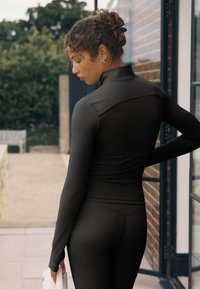 Zwart sporttopje met lange mouwen en hoge kraag, gecombineerd met bijpassende nauwsluitende legging. Gladde, rekbare stof en minimale stiksel details.