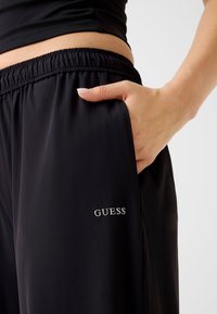 Pantalon de sport noir avec taille élastique, poches latérales et logo blanc "GUESS" sur la jambe gauche. Matière lisse et coupe ajustée.