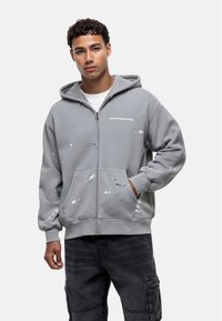 Felpa grigia con zip e cappuccio, caratterizzata da schizzi di vernice astratti e una tasca frontale. Testo sul petto: "GOODFORNOTHING." Tessuto misto cotone.