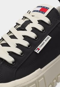 Μαύρο αθλητικό παπούτσι Tommy Jeans με χοντρές λευκές κορδόνια, ετικέτα με λογότυπο στο πλάι, χοντρή κρεμ σόλα και ετικέτα με λογότυπο στη γλώσσα.