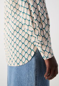 Long-sleeve shirt made of fabric featuring a cream background with a teal and beige geometric pattern. Buttoned cuff detail visible.
