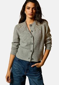 Cardigan grigio lavorato a maglia con scollatura rotonda, cinque bottoni e dettagli floreali decorativi sulle spalle. Texture morbida e vestibilità casual.