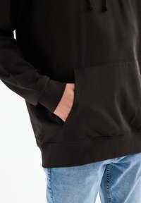 Sudadera negra con capucha y bolsillo frontal; tela suave con puños y dobladillo acanalados; usada con jeans de color azul claro.