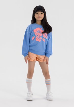 Sweatshirt bleu avec un motif floral rose, shorts froncés orange, baskets blanches et chaussettes montantes rayées. Tenue d'enfants simple et décontractée.