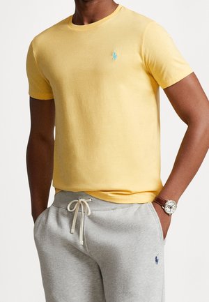 Mand iført gul kortærmet T-shirt og lysegrå joggingbukser med snor, hænderne i lommerne og brun armbåndsur på venstre håndled.