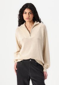 Beige strikket genser med halv glidelås i kragen, med en teksturert åpen vevemønster og ribbede mansjetter, sammen med svarte jeans.