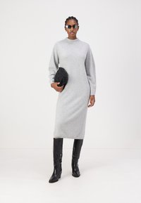 Robe midi en tricot grise avec un col montant et des manches longues, accompagnée d'une pochette texturée noire et de bottes noires jusqu'au genou.
