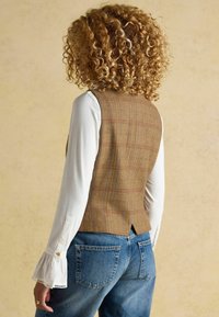 Hnedý károvaný vest s upraveným strihom, s rozdeleným chrbtom, vrstvený nad bielou blúzkou s dlhým rukávom, skombinovaný s modrými džínami.