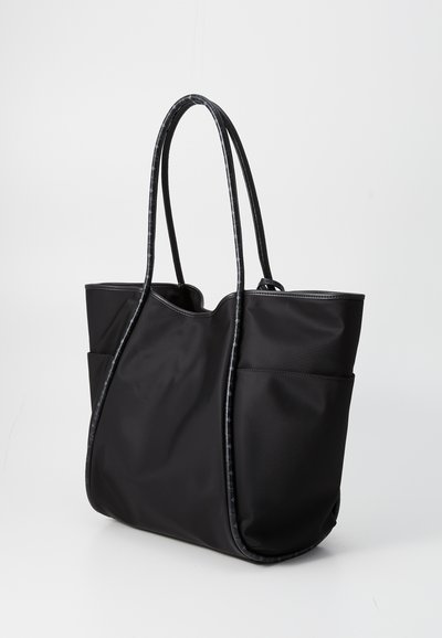 Bolsa tote preta com duas alças longas e dois bolsos laterais exteriores, de pé num fundo branco.
