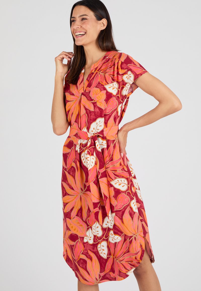 Robe florale présentant un fond rouge vif avec des motifs de feuilles roses et blanches, des manches courtes, un décolleté en V et une ceinture à la taille.