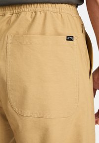 Shorts caqui con un bolsillo trasero con bordes cosidos y una pequeña etiqueta de marca. Cintura elástica y material de algodón ligero.