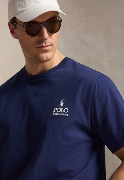 Man wearing a navy blue Polo Ralph Lauren t-shirt, dark sunglasses, and a white cap, looking slightly downward to the side.
