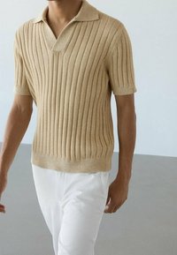 Polo en tricot beige avec texture côtelée verticale, manches courtes et col plat, associé à un pantalon blanc.