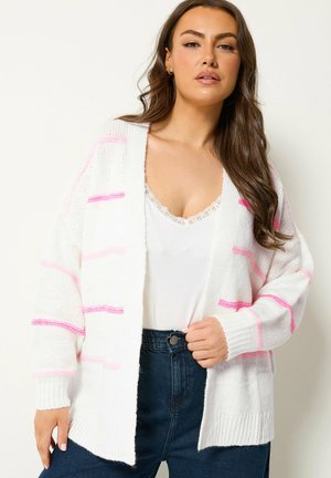 Kvinde iført hvid cardigan med pink striber over hvid bluse med blonder og højtaljede mørke jeans, poserer mod en ensfarvet baggrund.