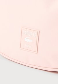 Rosa stoff med et sydd rektangulært lapp som viser Lacoste-logoen, krokodilleikonet og ordet «Paris» i hvitt.