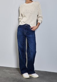 Pull tricoté beige clair avec un col rond, associé à un jean à jambes larges bleu foncé. Le mannequin porte des baskets blanches. Texture lisse, design décontracté.