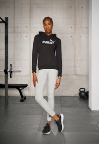 Mustaga kapuutsiga dressipluus valge Puma logo, hallid retuusid ja mustad spordijalatsid. Keskkond on spordisaal, kus on treeningvarustus.