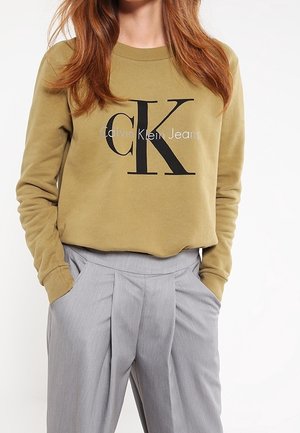 Sudadera verde oliva con el logotipo y el texto "CK" en negro en el pecho. Combinada con pantalones de traje plisados en gris claro. Tejido de mezcla de algodón, mangas largas.