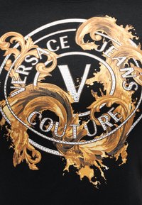 Μαύρο ύφασμα με μεγάλο κυκλικό λογότυπο που φέρει το κείμενο "Versace Jeans Couture" και περίτεχνους χρυσούς και μπρούντζινους μπαρόκ στροβίλους.