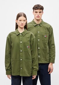 Μπουφάν από βαμβάκι σε ελιά πράσινο με κουμπιά στο μπροστινό μέρος, τσέπες στο στήθος και δύο κάτω τσέπες, με πολύχρωμο σήμα λογότυπο στην αριστερή πλευρά.