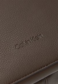 Brun strukturert lærflate med et preget "Calvin Klein"-logo, med jevn stikking langs kantene.