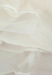 Mehak bel prosojen material z nežnimi vezenimi robovi in fino mrežasto teksturo, plasten čez gladko belo tkanino.