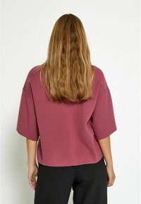 Loose-fit, short-sleeve top in mauve fabric. Soft texture with a rounded hemline, paired with black trousers. No visible patterns or hardware.