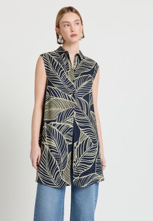 Femme portant une longue chemise sans manches avec un imprimé feuilles tropicales bleu marine et beige, associée à un jean bleu et de grandes boucles d'oreilles florales.