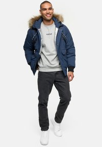 Marineblå pufferjakke med en pelsforet hætte, sølvfarvet inderside, frontlommer, parret med en grå sweatshirt og sorte jeans, afsluttet med hvide sneakers.