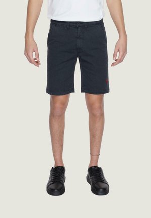 Pantalones cortos de algodón negros con un corte ajustado, trabillas para cinturón, bolsillos frontales y un logo bordado en rojo en el muslo izquierdo. Llevados con zapatillas negras.