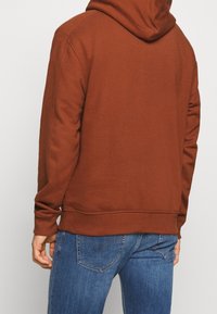 Persona che indossa una felpa color ruggine e jeans in denim blu, vista da dietro, in piedi contro uno sfondo chiaro e uniforme.