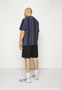 Camicia a maniche corte a righe navy, pantaloni corti neri, sneakers bianche e navy, logo visibile sui pantaloni corti, in piedi contro uno sfondo neutro.