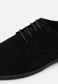 Scarpa da dress uomo in suede nero con punta rotonda, allacciatura chiusa e cuciture visibili lungo il lato e la suola.