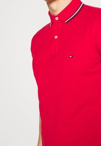 Rotes Poloshirt aus strukturiertem Baumwollstoff, mit einer Knopfleiste und einem kontrastierenden gestreiften Kragen. Kleines Logo-Detail auf der Brust.