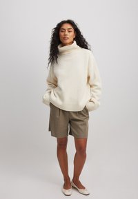 Kreemas krāsas pārmērīgs džemperis ar ribotu augstu apkakli un aprocēm, apvienots ar olīvzaļiem šortiem un gaišām kurpēm.