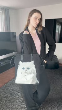 Fekete szatyor részletes ábrázolással egy fehér cicáról kék szemmel. A táskának sima fogantyúi és puha textúrája van.