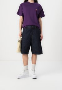 Persoon in een paarse T-shirt, donkerblauwe knie-lange shorts, witte hoge sneakers, witte sokken en met een beige tas in de hand.