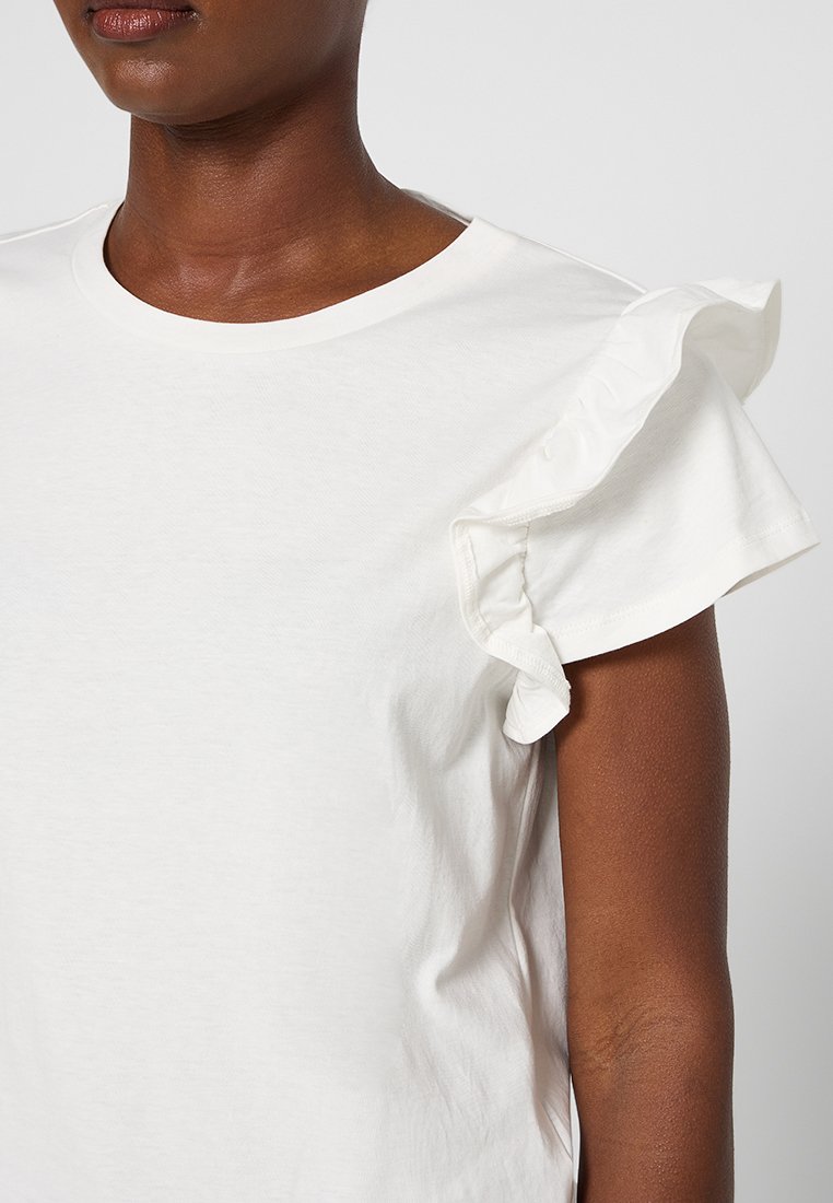 Eine Frau trägt ein weißes kurzärmliges Shirt mit Rüschendetails an einem Ärmel, gezeigt von Hals bis Oberarm vor einem schlichten Hintergrund.