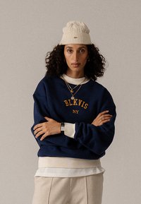 Tengerészkék pulóver narancssárga „BLKVIS NY” felirattal, fehér inggel rétegezve, bézs nadrággal és törtfehér kötött sapkával párosítva.