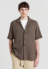 Camicetta marrone a maniche corte con texture, chiusura a bottoni, colletto classico e bottoni neri, indossata sopra una camicia bianca.