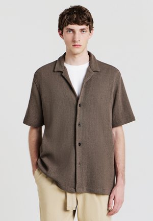 Chemise en tissu texturé marron à manches courtes avec une fermeture à boutons, un col classique et des boutons noirs, portée par-dessus une chemise blanche.