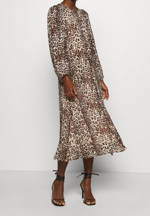 Vrouw draagt een langwerpige, knielange jurk met luipaardprint en zwarte enkellaarzen met hoge hakken en enkelbandjes, staand tegen een effen achtergrond.